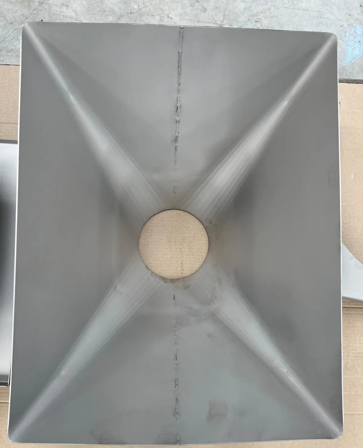 Vue plongeante dans une trémie inox : quatre faces qui convergent vers l'orifice de sortie circulaire, cordons de soudure visibles.
