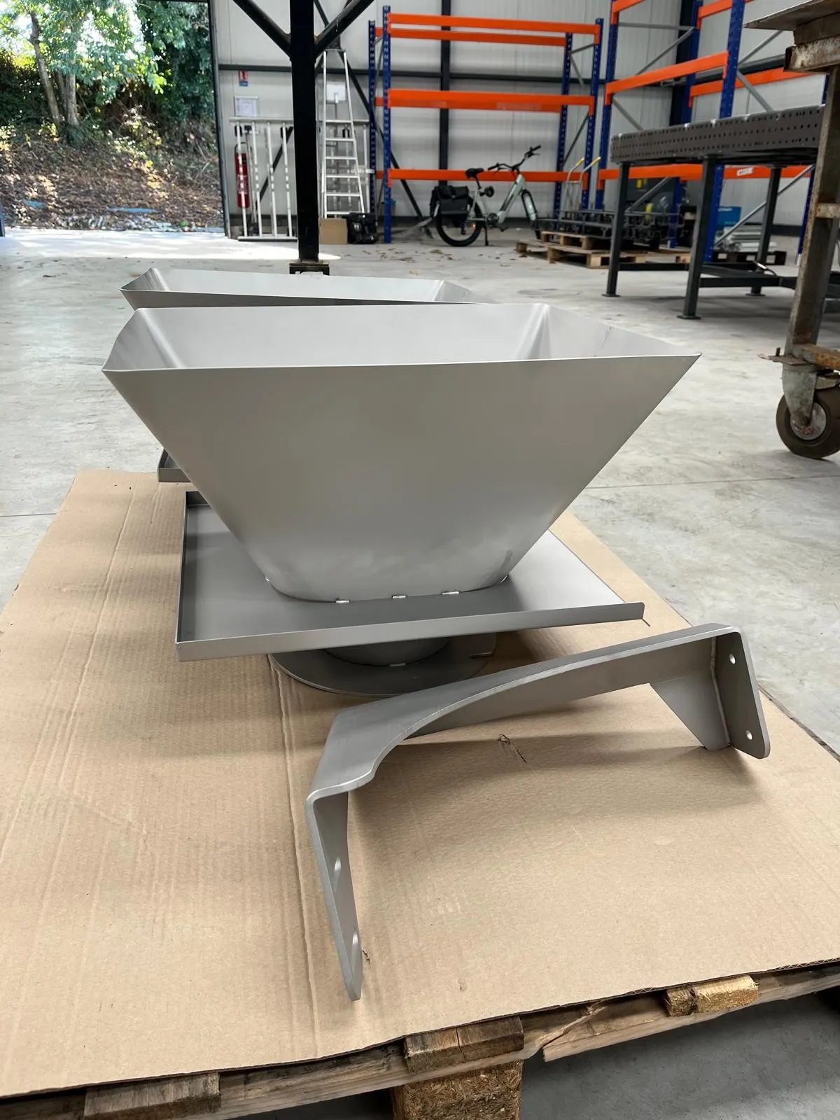 Deux trémies en inox posées sur une palette dans l'atelier, avec leurs platines de fixation et un support plié au premier plan.